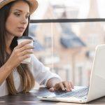 Working outdoors. Beautiful young woman in funky hat working on laptop and smiling while sitting outdoors